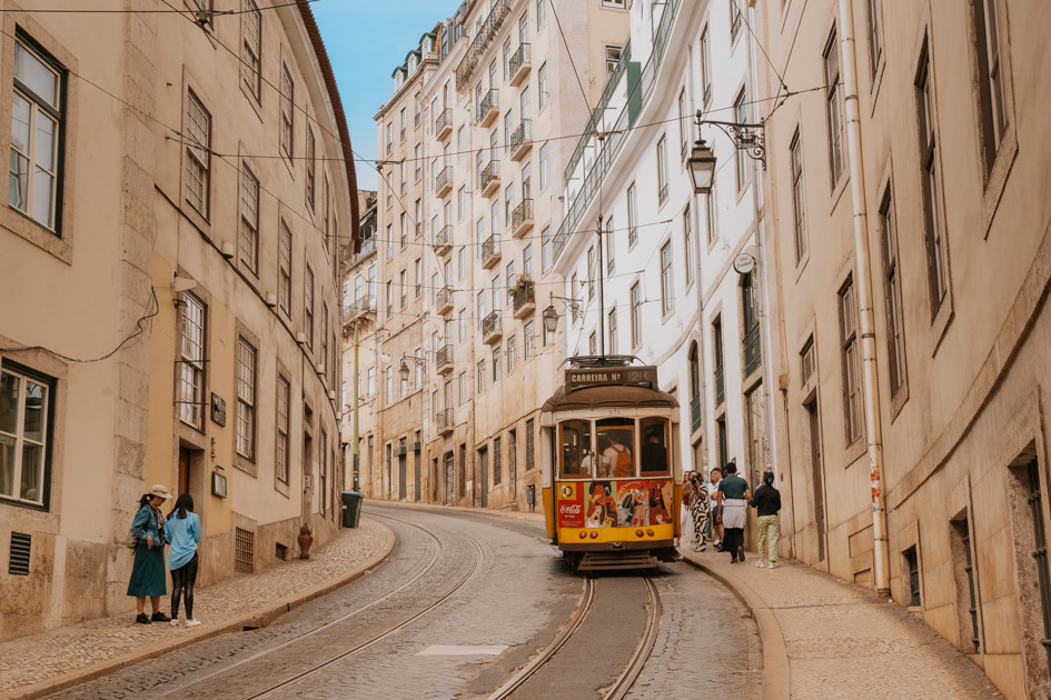 a historic red and yellow tram rides along a track in the middle of tall white buildings in downtown Lisbon Portugal