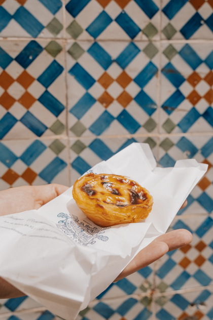 a close up of a creamy custard tart with toasted top on a white bag held by someone's hand against a colourful tiled wall in Belem Lisbon