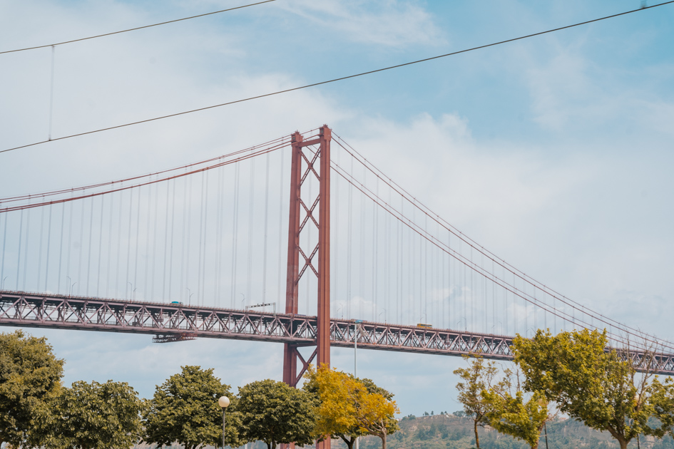 a red painted suspension bridge in the distance on a clear blue day in Lisbon with trees underneath