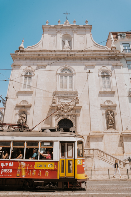 a red and yellow tram runs in front of a white stone church with clear blue sky on a Lisbon itinerary 2 days