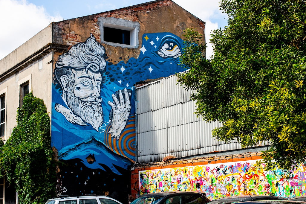 large scale graffiti art on the side of a building with man's fae and blue waves at LX Factory in Lisbon
