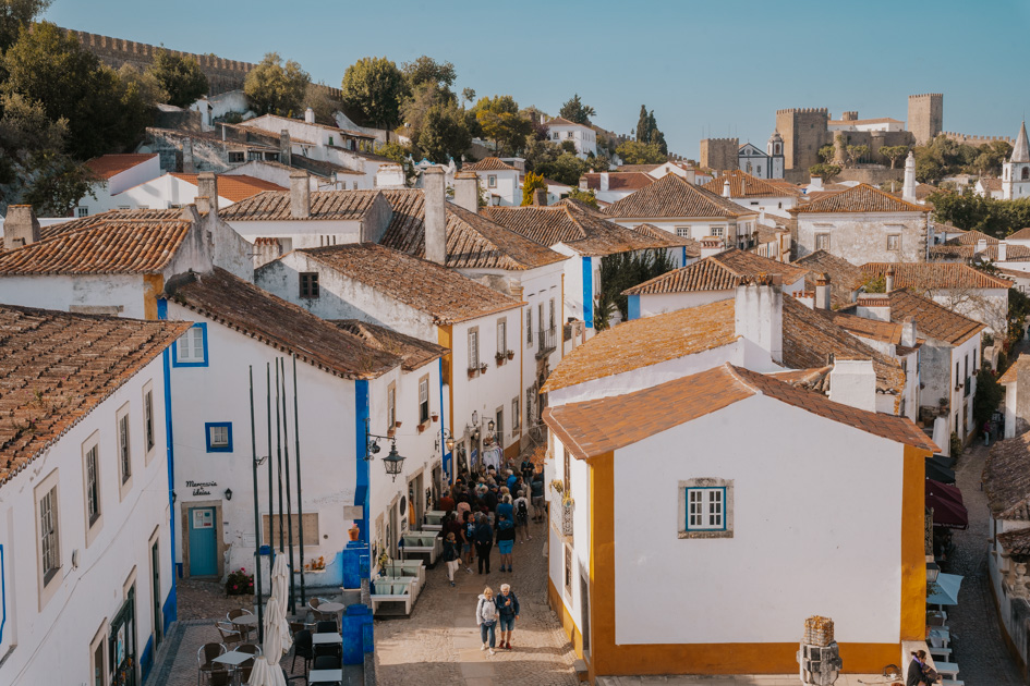 brown tile rooftops with white, blue and orange buidlings lie beneath a medieval wall in Obidos, one of the best historical day trips from Lisbon Portugal