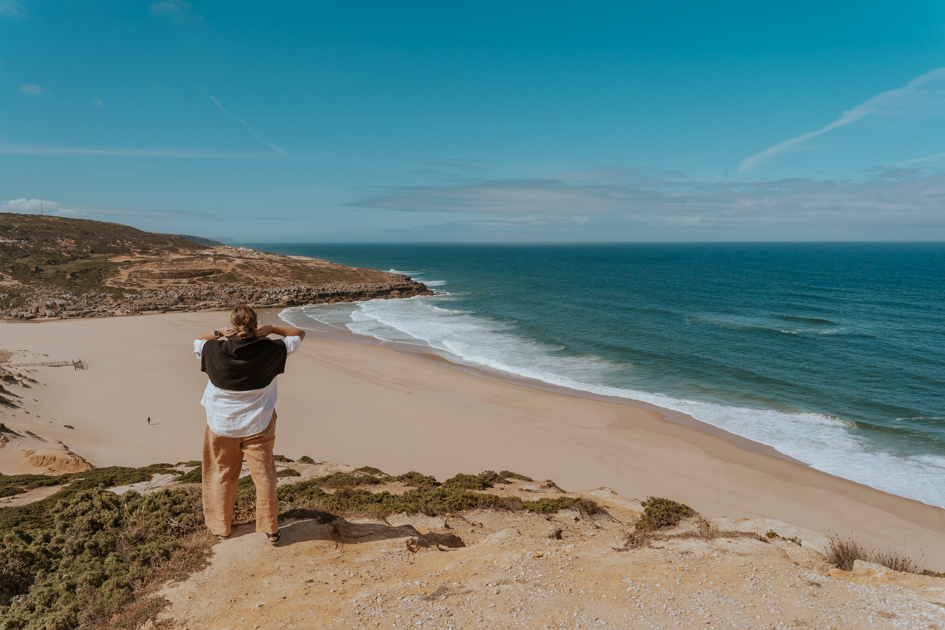 Haley Blackall wearing beige pants, white and black top stands on a clifftop overlooking the famous surf beaches of Ericeira with the blue Atlantic Ocean in the background from a Lisbon day tour