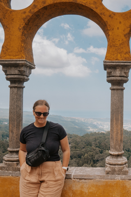 Haley Blackall smiles wearing beige pants and black t shirt infront of a yellow arch with panoramic views beyond in Sintra day tour from Lisbon