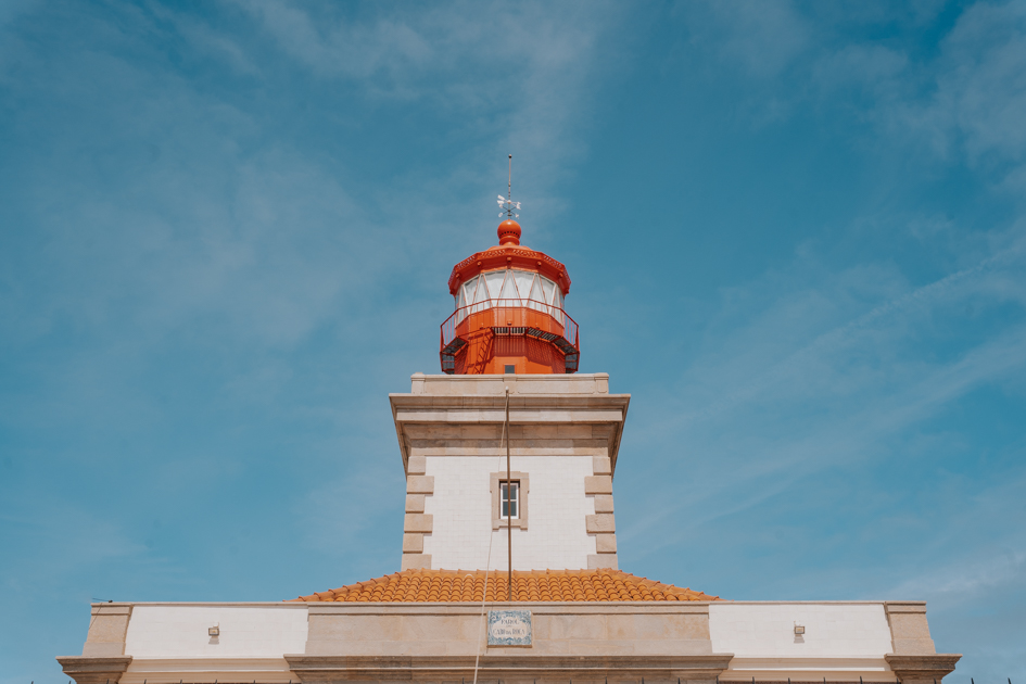 a red, white and orange lighthouse stands tall with a blue sky background at Cabo da Roca from Lisbon