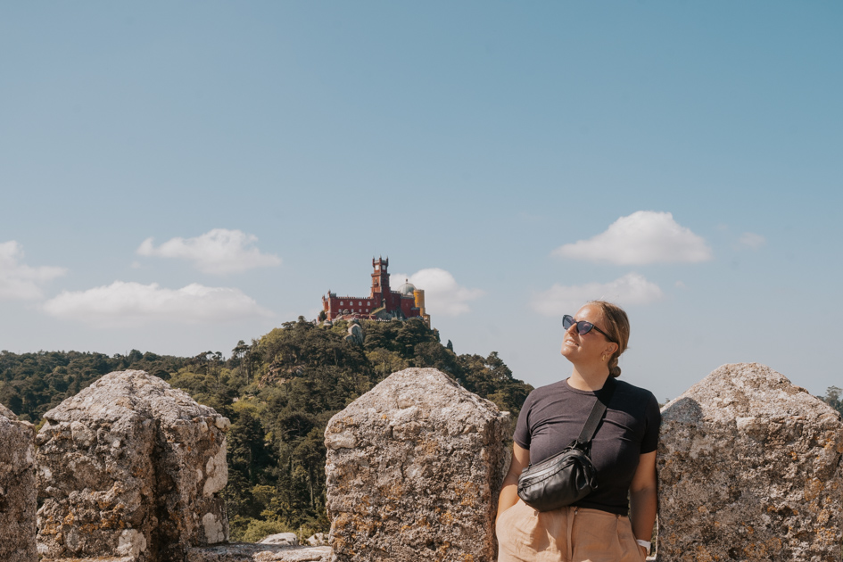 Haley Blackall wearing a black shirt and purse smiles next to a stone wall looking out at Pena Palace in Sintra, the perfect day trip from Lisbon
