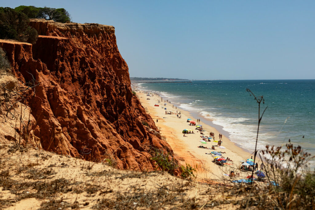 expansive golden sandy beach sits at the bottom of a red rugged cliffside with ocean and blue sky in one of the best places to stay in Algarve Portugal