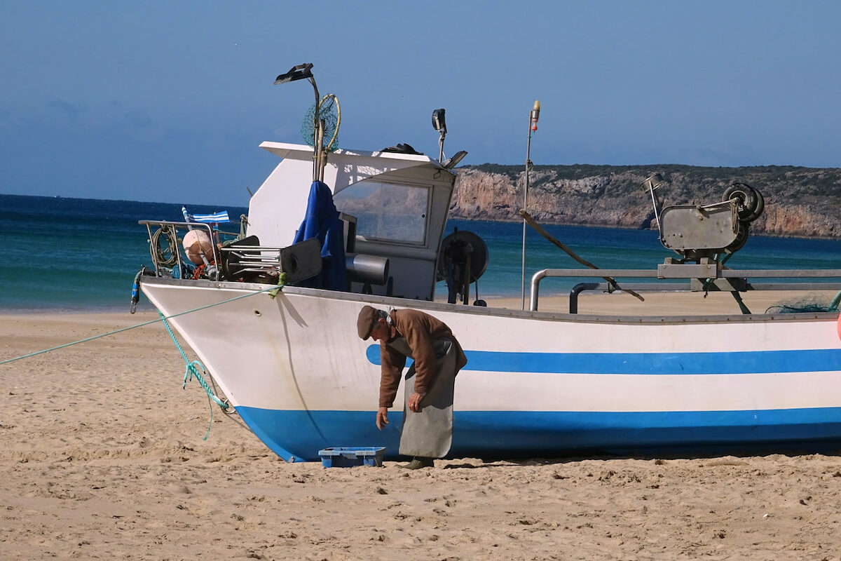 a fisherman bends over in front of a blue and white striped boat on a sandy beach in Algarve best places to stay