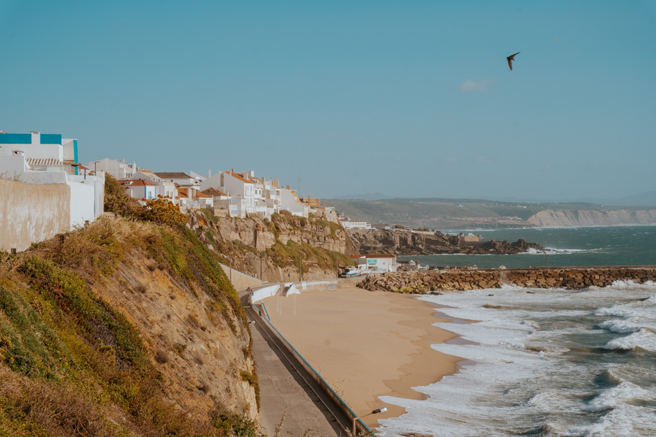 a sandy beach lies at the bottom of a rugged green cliff face on the left with a coastal town sitting on the top on a blue day on the Silver Coast Portugal