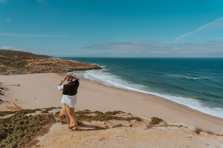 Haley Blackall wearing beige white and black stands on the edge of a lookout over an expansive sandy beach and Atlantic Ocean in the distance in Ericeira, where to stay on Portugal's Silver Coast for surfing