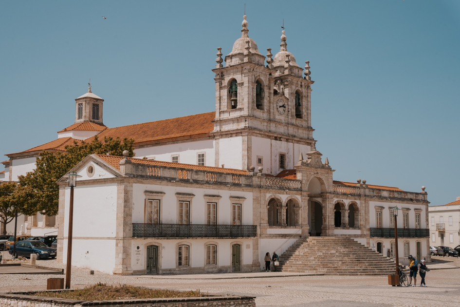 a gothic church with grey stone, white stone and red tiled roof lies behind a plaza with trees on a blue day in Nazare, one of the best places to stay on Portugal Silver Coast for big wave surfers