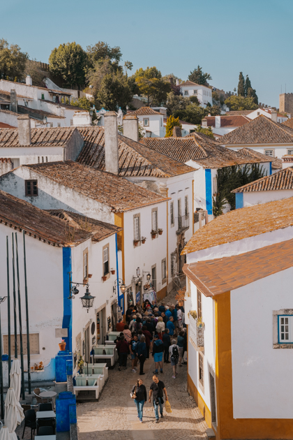 overlooking an charming street full of people lined with white, yellow, blue buildings on a sunny day in Obidos on the Silver Coast of Portugal