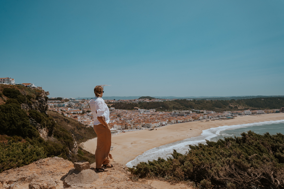 Haley Blackall stands on a outcrop looking over Nazare's town centre and white sandy beach on a clear day along the West Coast of Portugal