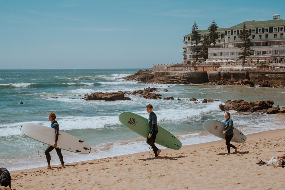 three beginner surfers carry their surfboards on a sandy beach in Ericeira with a multi-story hotel in the distance on a rocky outcrop in Portugal
