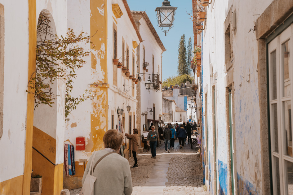 an elderly woman in a beige sweater walks down a cobblestone street lined with white buildings in Obidos Portugal town