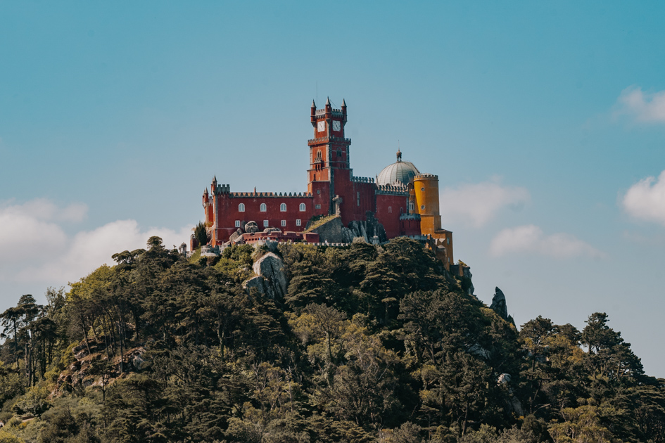 a red and yellow castle sits on top of a green hill on a blue day in Sintra one of the most romantic places to stay on Portugal Silver Coast