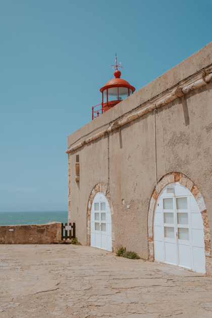 a grey stone fortress with white arched doors and red lighthouse with ocean in the distance in Nazare Portugal coastal town
