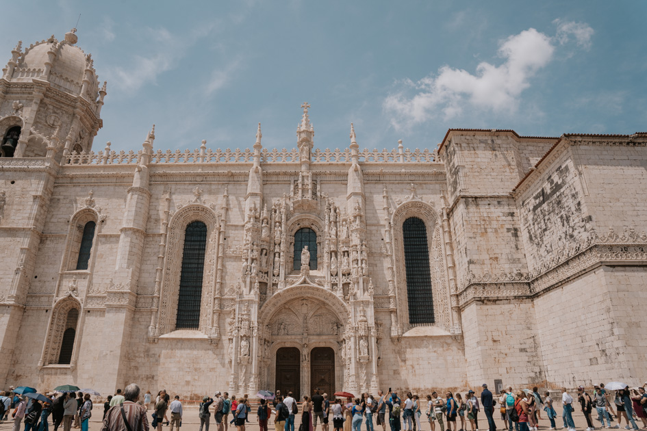 Looking up at Jeronimos Monastery, a Lisbon attraction with a line of tourists in the foreground on a blue day in Portugal