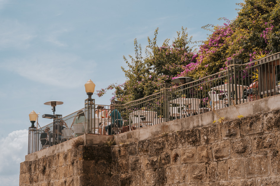 a panoramic patio on top of a grey stone wall with thin iron fence and blooming greenery on a blue day in Lisbon what to do