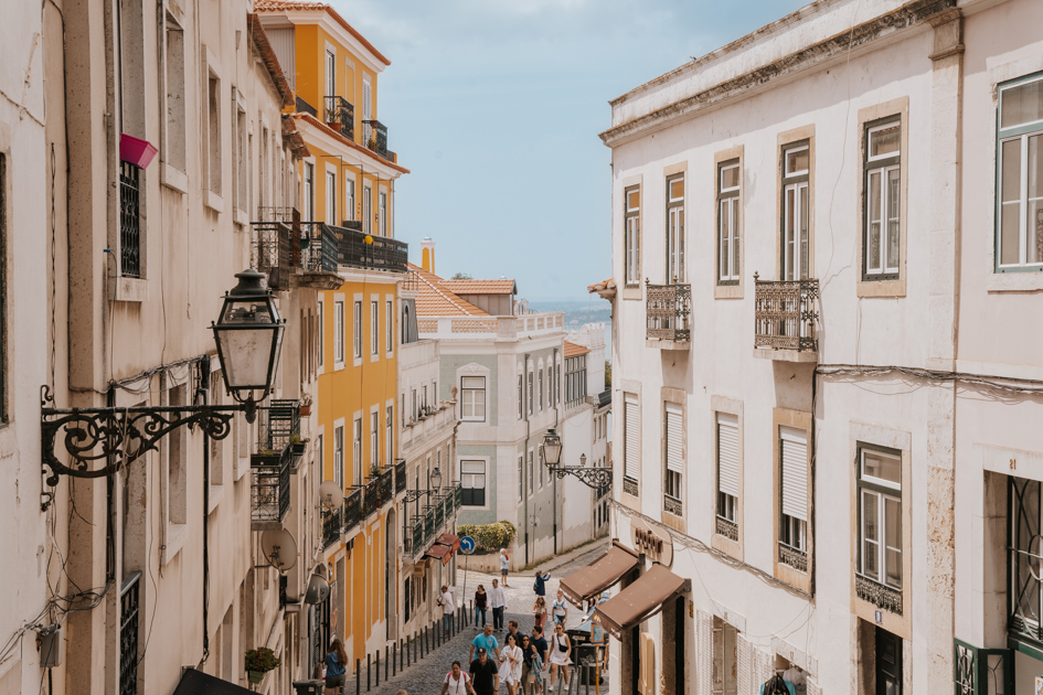 tourists walk up and down a steep street amongst yellow and white buildings in Alfama Lisbon Portugal