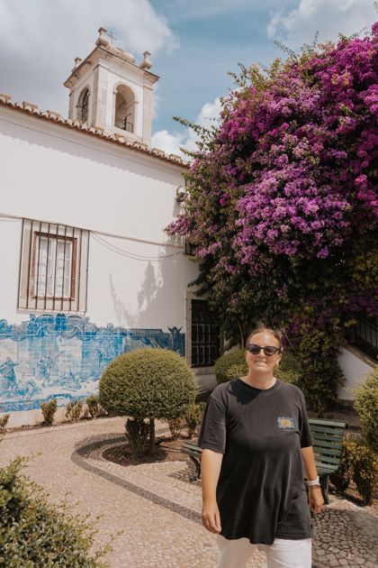 Haley Blackall wearing a black t shirt smiles at the camera while walking in a courtyard with purple flowers and blue tiled wall as a first timer in Lisbon Portugal