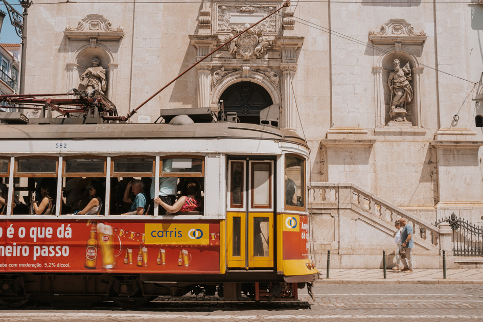 a red and yellow old tram drives in front of a historic white stone building in downtown Lisbon Portugal