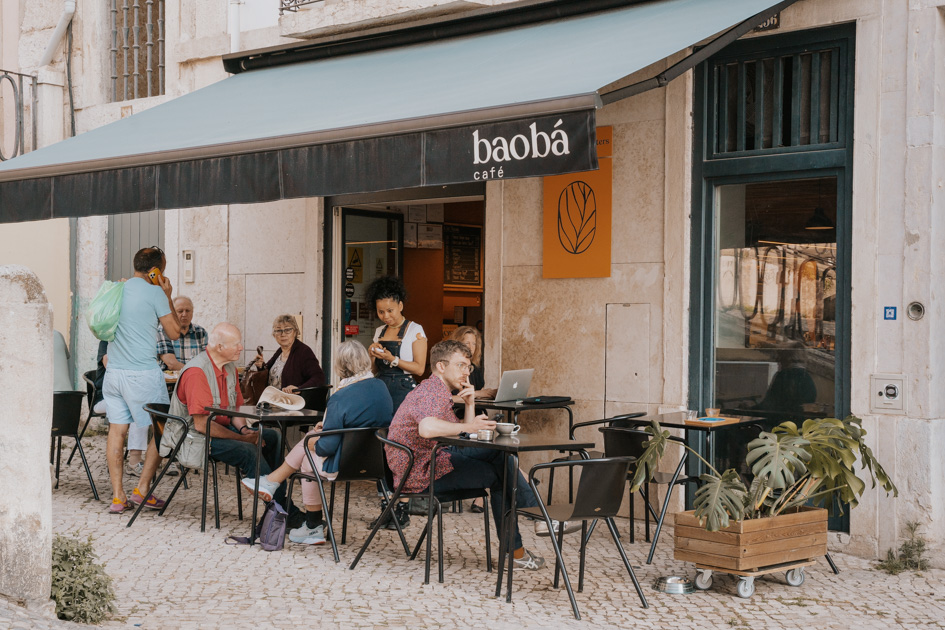 people sit on a streetside patio under a black awning that says 'baoba cafe' in Chiado, Lisbon travel tips