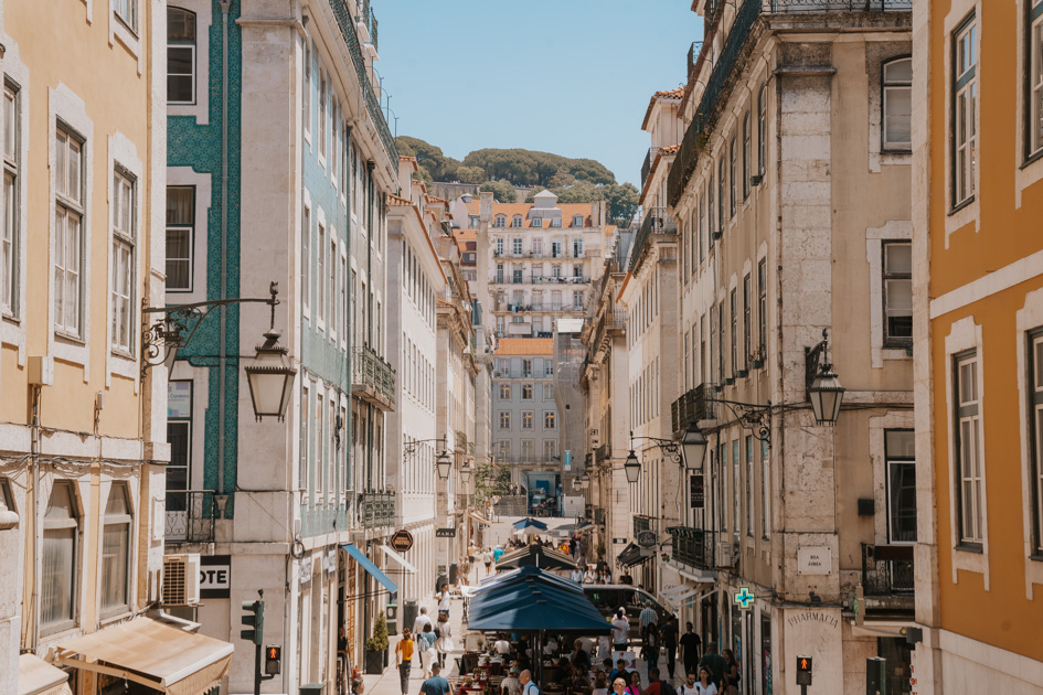 pastel coloured buildings line a bustling streets capped off with the walls of the Sao Jorge castle in downtown Lisbon, Portugal
