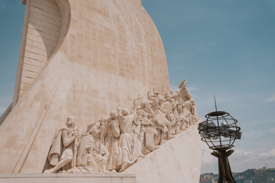 a close up perspective of stone people climbing upwards on the side of a large curve shaped monument with black iron globe on a clear day on an itinerary for Lisbon Portugal