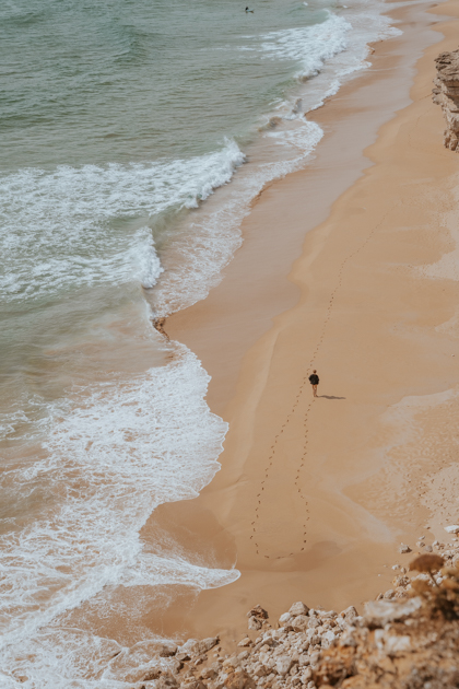 person in black walks along an desolate sandy beach with waves crashing in Sagres best town in Algarve
