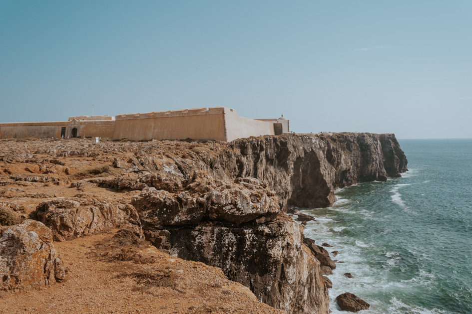 Low concrete walls of the Sagres Fortress sit at the edge of a rugged coastline on a sunny day in one of the best areas to stay in Algarve Portugal