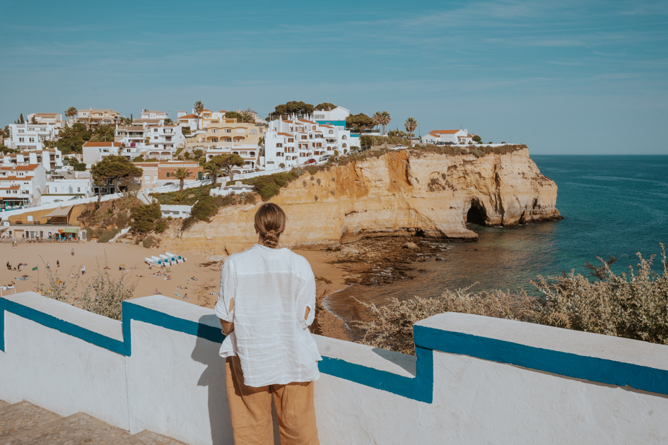 a woman wearing white and beige stands before a blue and white concrete wall overlooking Carvoeiro which is where to stay in Algarve Portugal