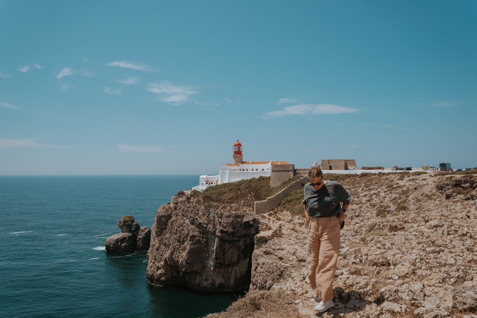 a girl wearing black and beige walks on the edge of a coastal cliff with a white and red lighthouse in the distance on a sunny day in one of the best towns in Algarve Sagres