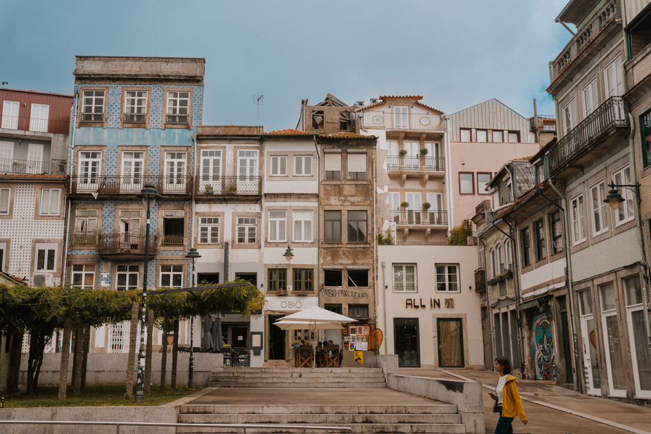 concrete plaza with tall thin buildings behind and a woman walking with a yellow jacket in Bonfim the best place to stay in Porto for budget travellers