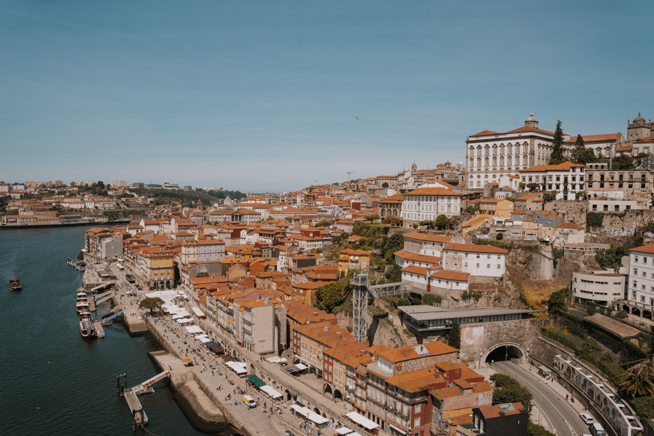 red roofed historic buildings line a hillside with a bustling riverfront promenade and expansive river on a sunny day is where to stay in Porto for couples