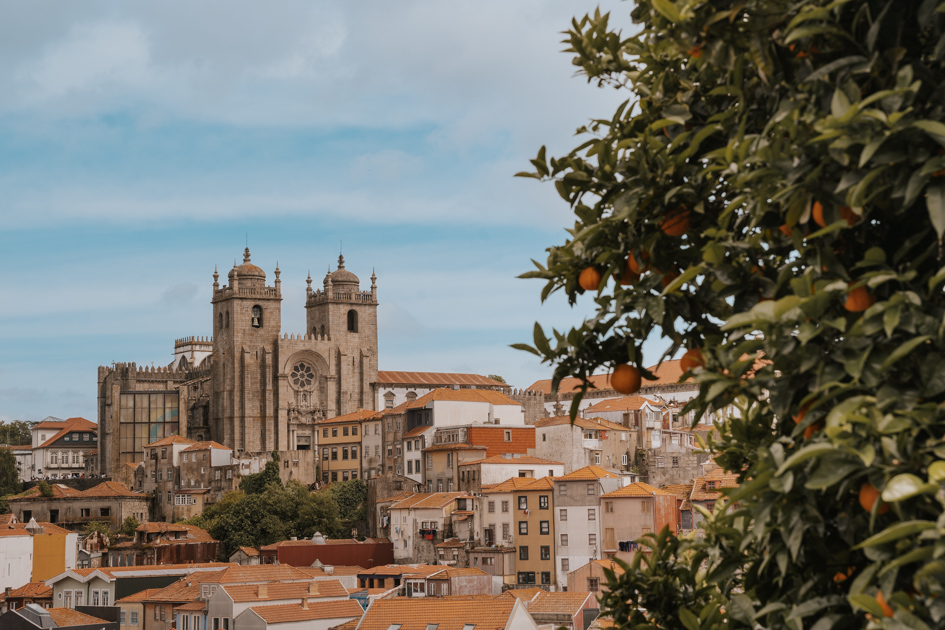 Se Cathedral sits on the top of a hill with red roofed buildings below and orange tree in the foreground on a sunny day in Baixa one of the best locations to stay in Porto