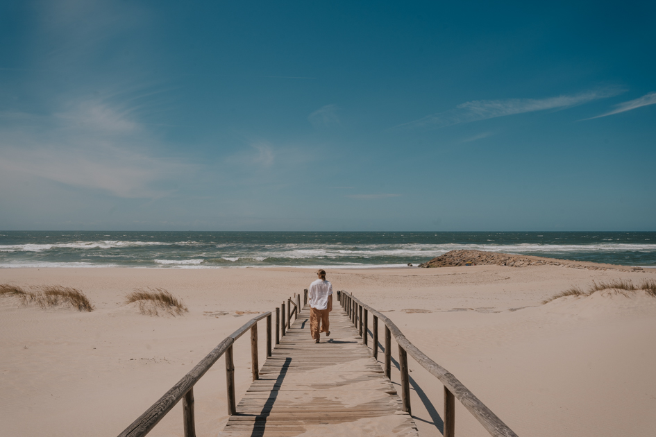 Haley Blackall wearing beige pants and flowy white top walks along a wood boardwalk out to an expansive sandy beach and ocean beyond on a blue day in Costa Nova, where to stay on Portugal's Silver Coast for beaches
