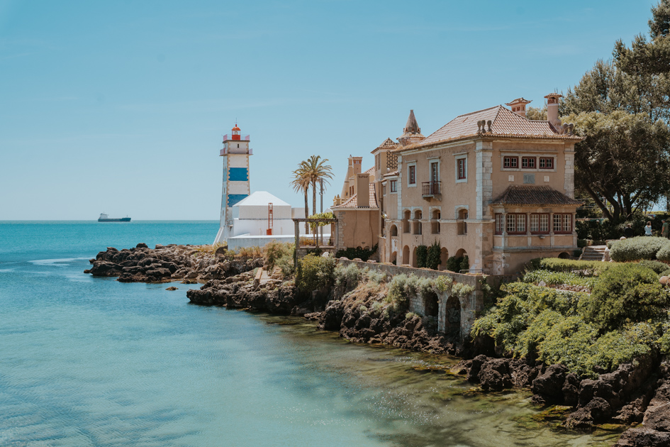 subdued pink building and blue and white striped lighthouse sit on a rocky peninsula with greenery and azure blue waters in Cascais one of the best places to stay on the Silver Coast of Portugal for luxury
